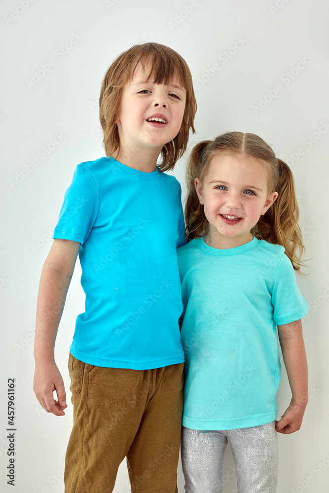 Cheerful toddler girl and preschooler boy in bright blue t shirt hugging and looking at camera while standing against white wall Cheerful toddler girl and preschooler boy in bright blue t shirt hugging and looking at camera while standing against white wall