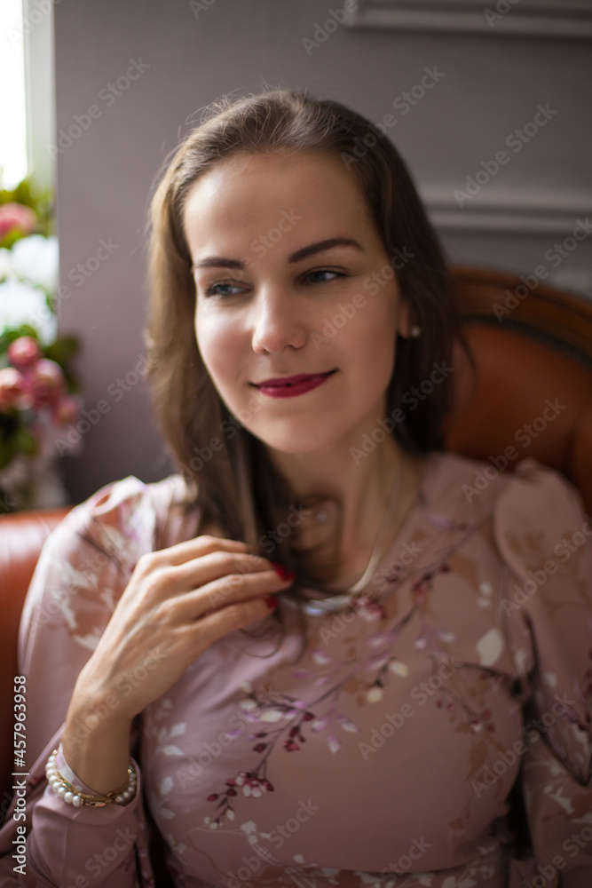 Naklejka premium Young beautiful woman in pink dress and stylish accessories sits on brown leather sofa on white wall background. Portrait of happy smiling girl with red lips resting at home and looking at camera. 