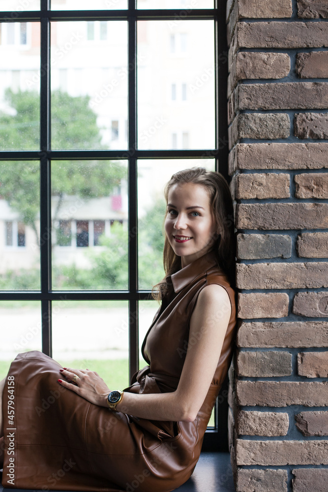 Beautiful young woman in brown leather dress resting on the windowsill. Portrait of happy smiling girl sits on the stained glass window at industrial loft wooden brick style interior. Leisure concept.