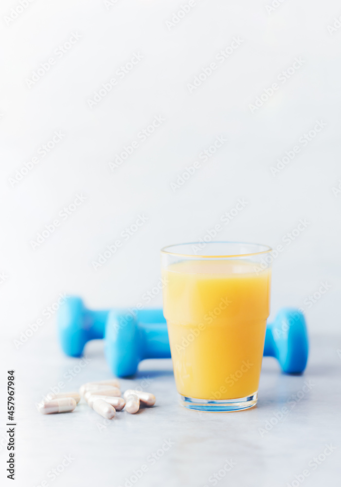 Glass of Orange Juice. Blue Dumbbells and Sport supplements ( Carnitine capsules ) in background. Stone background. Close up. Glass of Orange Juice. Blue Dumbbells and Sport supplements ( Carnitine capsules ) in background. Stone background. Close up.