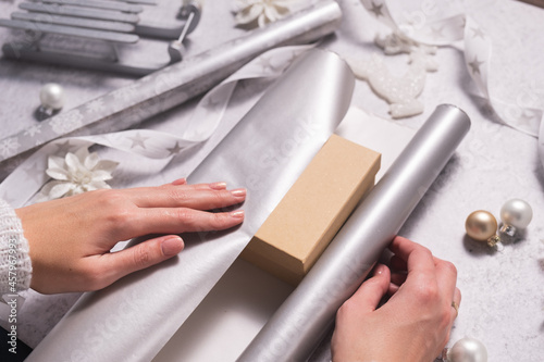 Women's hands are wrapping a Christmas present in silver wrapping paper.