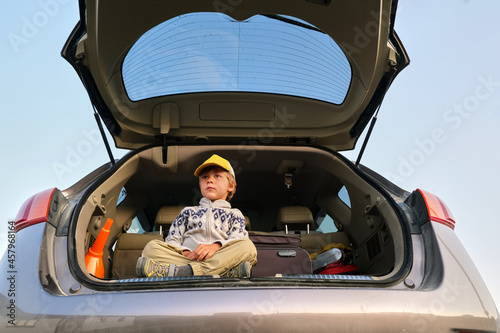 Low angle of dreamy little boy in hoodie and cap sitting with legs crossed in open trunk of modern automobile with suitcase before summer journey