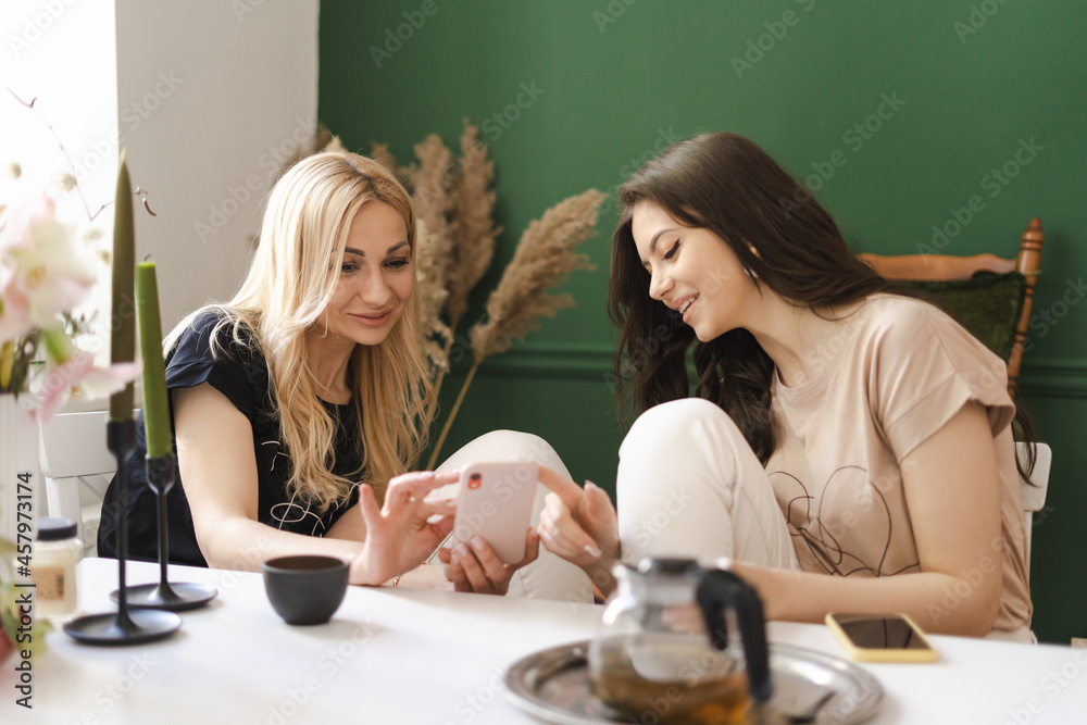 Photo of pleased caucasian mother and daughter using cellphone while having tea in modern kitchen Photo of pleased caucasian mother and daughter using cellphone while having tea in modern kitchen