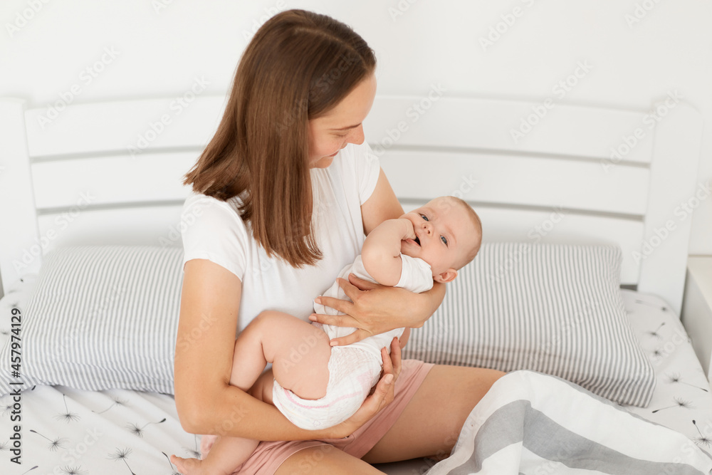 Dark haired female holding baby girl in hands, looking at her tiny daughter with great love, posing indoor sitting on bed with her lovely child, happy motherhood. Dark haired female holding baby girl in hands, looking at her tiny daughter with great love, posing indoor sitting on bed with her lovely child, happy motherhood.