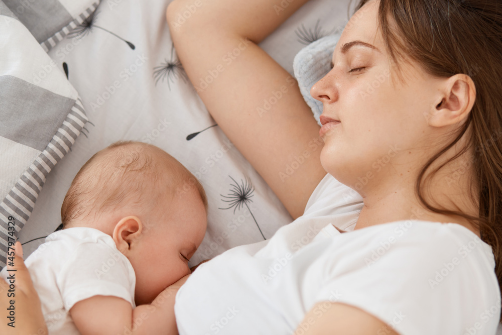Portrait of sleepy woman feeding breast her baby in the bed sleeping together, young adult female mommy wearing white casual t shirt, resting while her toddler kid sleeps. Portrait of sleepy woman feeding breast her baby in the bed sleeping together, young adult female mommy wearing white casual t shirt, resting while her toddler kid sleeps.