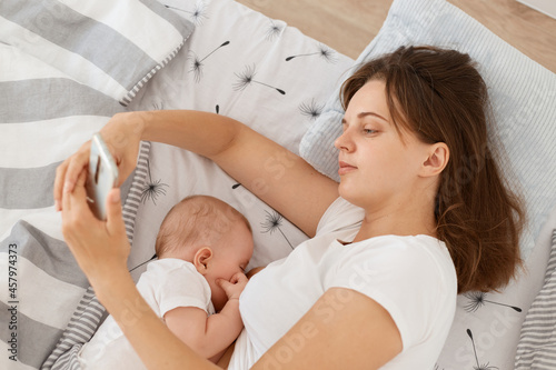 Good looking tired sleepy woman wearing white casual style t shirt using cell phone for checking networks or typing messages while breast feeding her infant baby, lying bed. photo