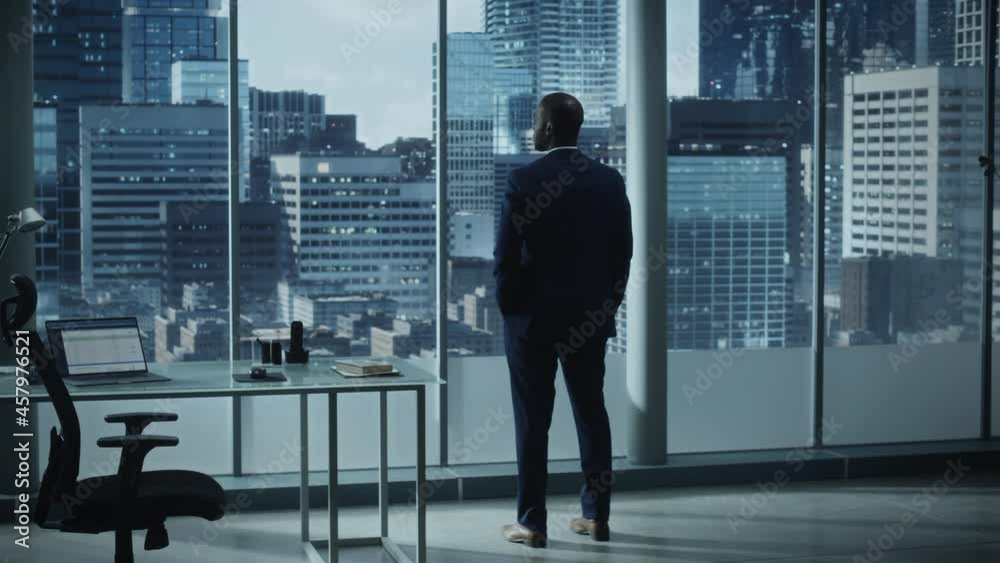 Thoughtful African-American Businessman in a Perfect Tailored Suit Standing in His Office Looking out of the Window on Big City. Successful Investment Manager Planning Strategy for e-Commerce Startup