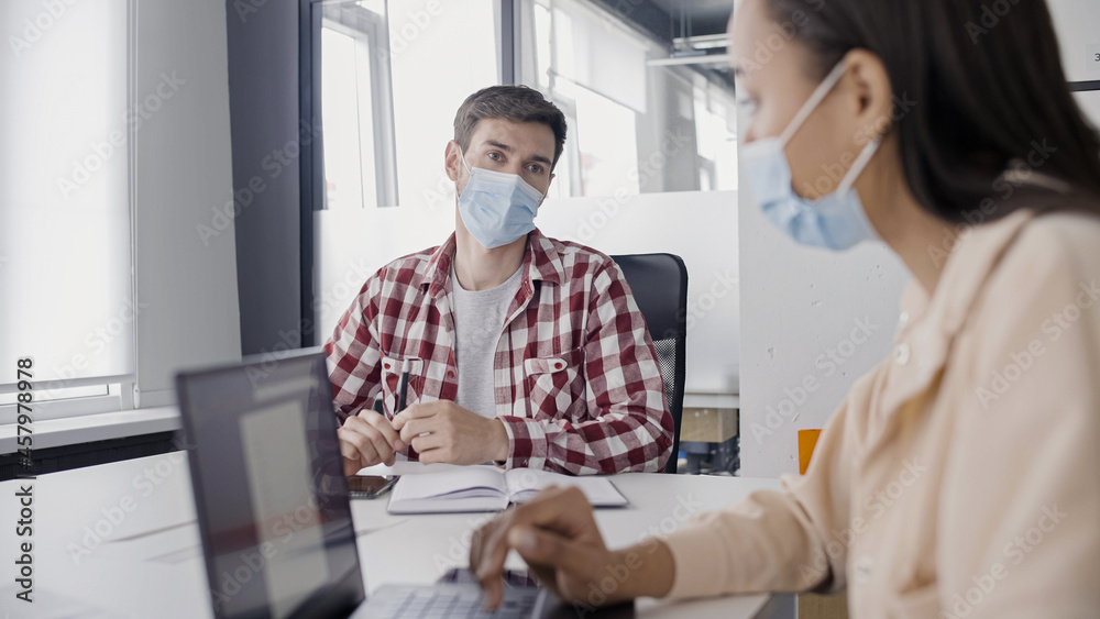 Personal assistant in mask typing on laptop, talking to businessman in office Personal assistant in mask typing on laptop, talking to businessman in office