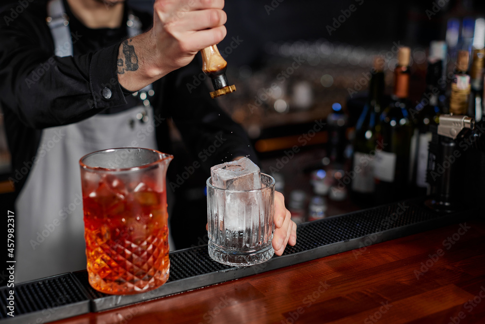 barman in apron pours old fashioned cocktail into glass