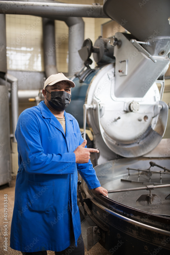 Portrait of workman standing next to coffee roasting machine. Interior of coffee production workshop with set up roasting equipment. Person in uniform and mask gesturing. Production, food concept