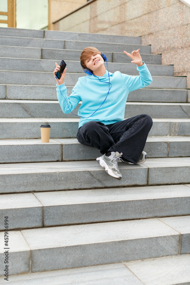 Vertical photo of a young hipster woman sitting on the stairs and listening to music through an online smartphone app and headphones. She is happy and free.
