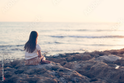 Sad lonely Caucasian child preteen girl sitting on stones by the sea, toning, negative emotions, emptiness and sadness