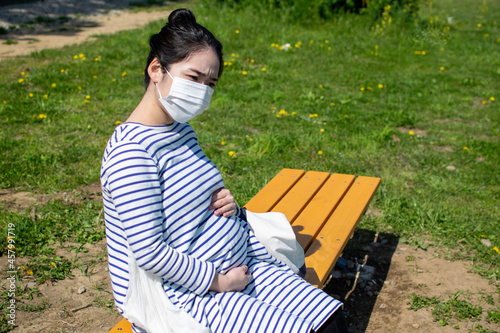 A young pregnant Asian woman sits on the outside bench and worries about her baby in her stomach