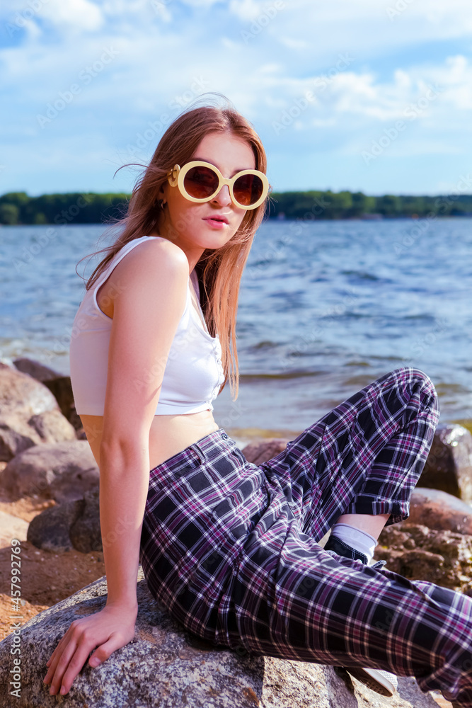 Caucasian Blond Female Girl in casual Clothing Posing on Coastal Stones on Sea Shoreline Outdoors At Sunny Day.