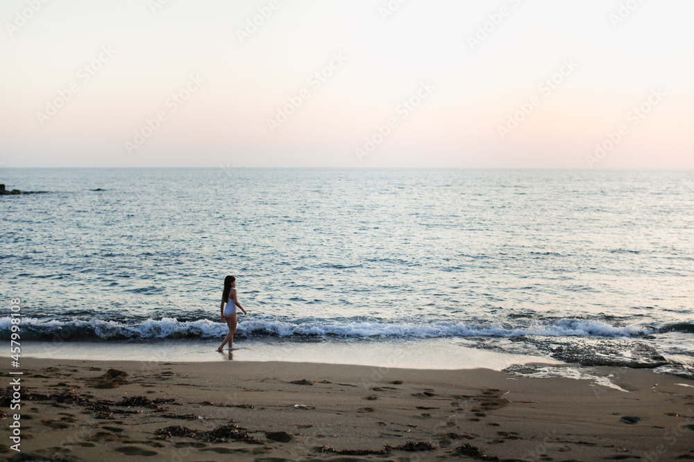 Cute caucasian girl in a striped swimsuit with long hair runs seashore on a deserted beach at a resort, evening and sunset