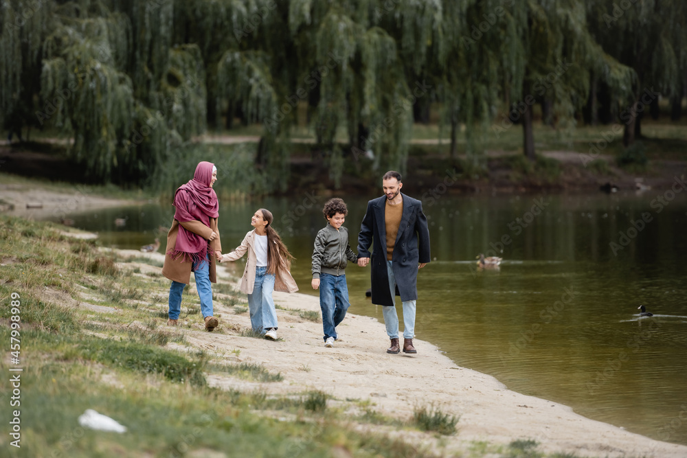 Fototapeta premium Positive arabian family holding hands while walking near lake