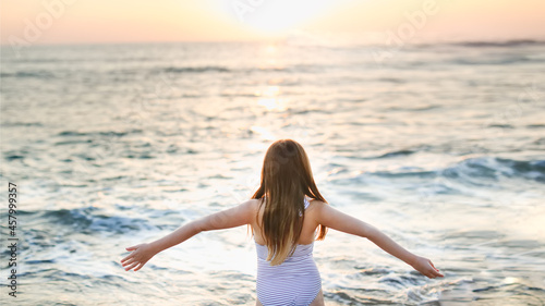 Cute caucasian child girl in a striped swimsuit with long hair with outstretched arms hugs the sea on an empty beach at a resort, evening and sunset © natalialeb