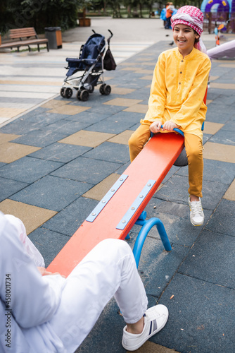 Fototapeta Naklejka Na Ścianę i Meble -  Smiling arabian boy in kufiya playing with blurred father on playground