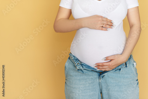 No face portrait of young pregnant woman in blue jeans and white T-shirt on the beige background.