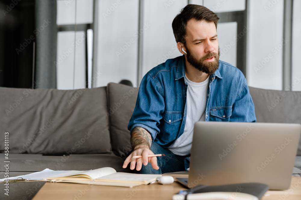 Bearded european man working with laptop while sitting on sofa Bearded european man working with laptop while sitting on sofa