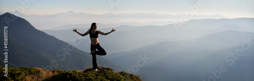 Panoramic view of fit young woman performing yoga pose on grassy hill with mountains and sky on background. Beautiful woman in sportswear practicing yoga outdoors. Copy space.
