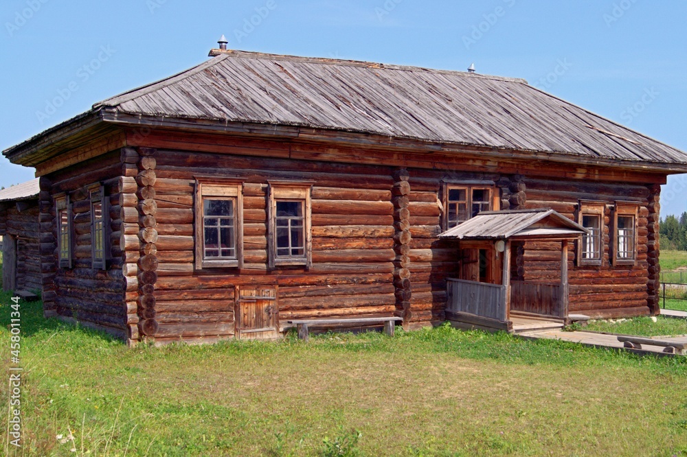 Wooden house made of logs on a clear summer day. The wooden house was built in the 19th century. Wooden house made of dark logs, blue sky, green grass in front of the house. Wooden house made of logs on a clear summer day. The wooden house was built in the 19th century. Wooden house made of dark logs, blue sky, green grass in front of the house.