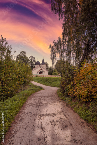 A road leading to the orthodox church of white bricks against a bright sunset sky. Russian village with traditional buildings on a summer evening.