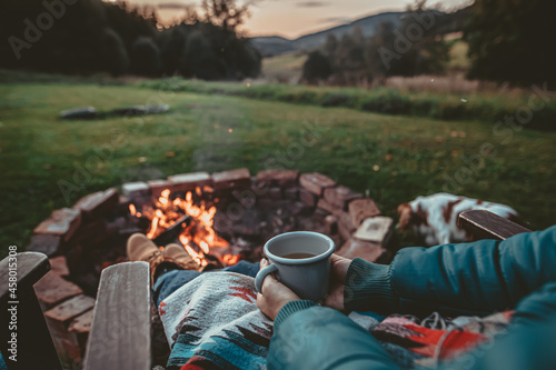 Unrecognizable Woman Enjoying Hot Tea From A Tin Cup In Campsite With Fire Pit. Girl In Folk Blanket By Burning Campfire with mountain landscape with evening sunset sky over the forest and hills.