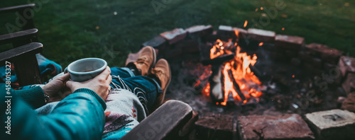 Unrecognizable Woman Enjoying Hot Tea From A Tin Cup In Campsite With Fire Pit. Girl In Folk Blanket By Burning Campfire with mountain landscape with evening sunset sky over the forest and hills.