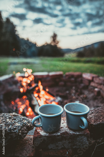 Campsite With Fire Pit and Two Tin Cups with hot tea. Burning Campfire with mountain landscape with evening sunset sky over the forest and hills.