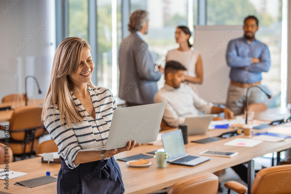 Smiling woman with a laptop in office. Portrait of a smiling confident young businesswoman holding laptop in hand looking at camera with colleague at background in office Smiling woman with a laptop in office. Portrait of a smiling confident young businesswoman holding laptop in hand looking at camera with colleague at background in office