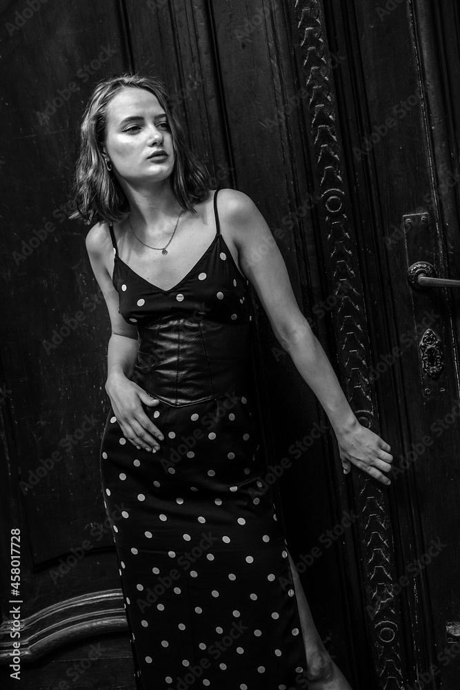 Black and white stylish portraits of a brunette young girl in an urban environment. Black summer dress with white polka dots, red lips and a slight melancholy