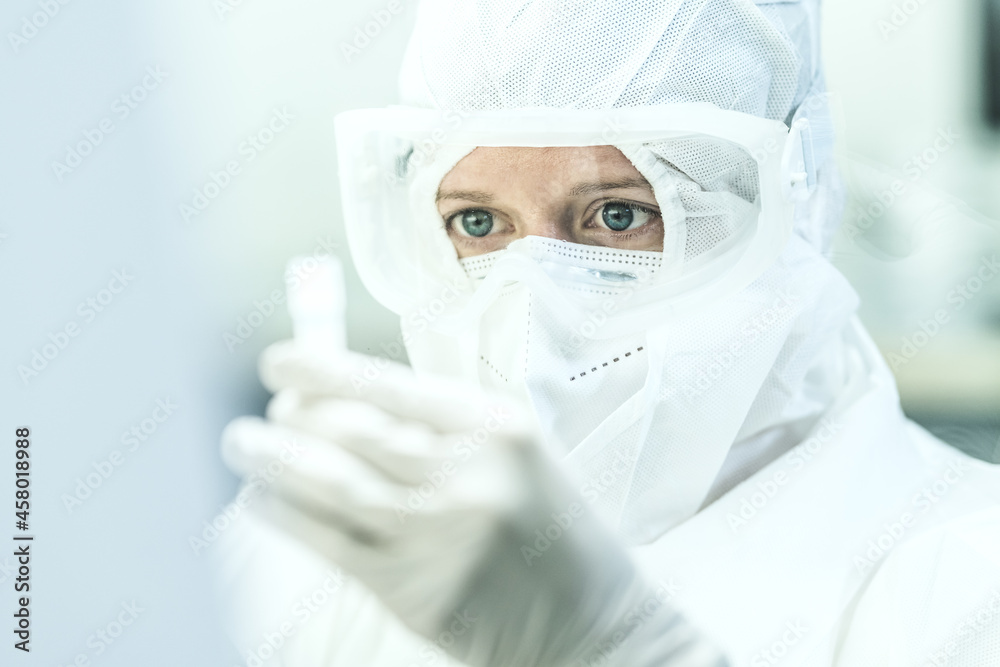 Close-up shot of a Caucasian female scientist in a protective suit, with glasses, face mask, and sterile gloves, looking worried at a test tube in her hand. Close-up shot of a Caucasian female scientist in a protective suit, with glasses, face mask, and sterile gloves, looking worried at a test tube in her hand.