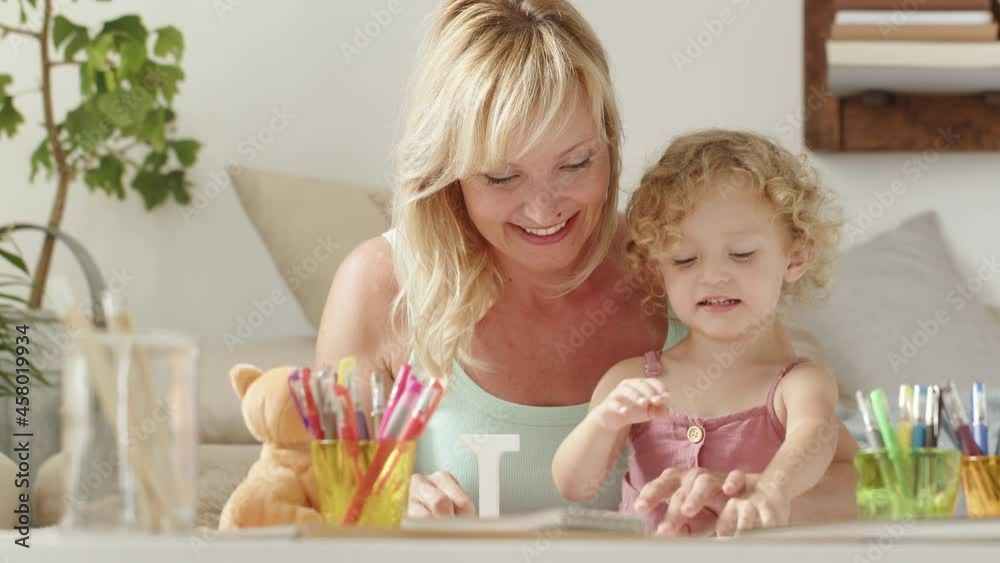 smiling mother smiles at curly haired blonde daughter child, preschool learning activity at home, concept of healthy growing, reads and writes the word love cheerfully playing in sunny living room
