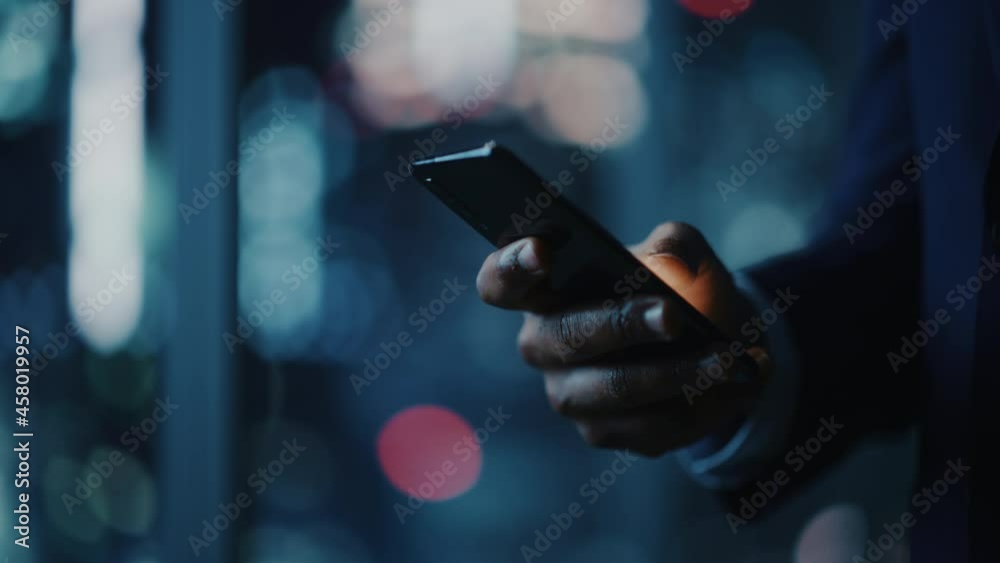 Night Office: Successful Black Businessman Wearing Suit Standing, Using Smartphone. CEO Browsing Internet, Managing Social Media Strategy in e-Commerce Software. Focus on Hand with Mobile Phone