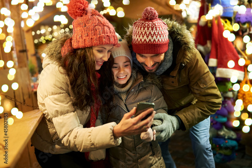 family, winter holidays and technology concept - happy mother, father and little daughter with smartphone at christmas market in evening