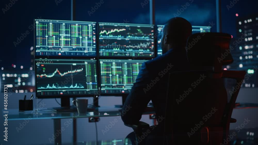 Hedge Fund Trader Working on a Computer with Multi-Monitor Workstation with Real-Time Stocks, Commodities and Exchange Market Charts. Businessman Works in Investment Bank Downtown Office at Night. Hedge Fund Trader Working on a Computer with Multi-Monitor Workstation with Real-Time Stocks, Commodities and Exchange Market Charts. Businessman Works in Investment Bank Downtown Office at Night.