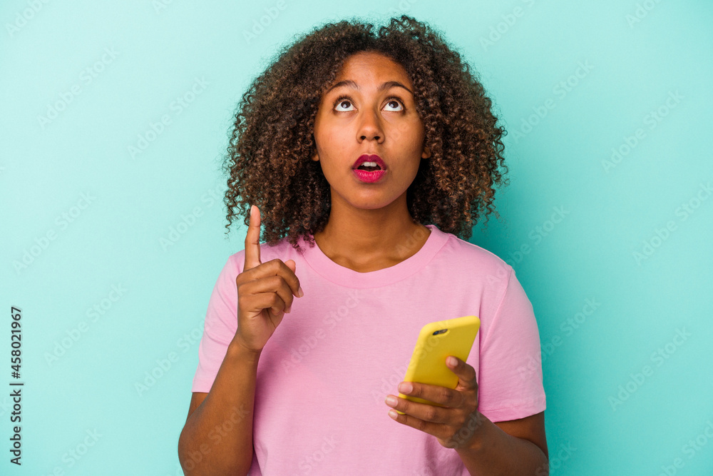 Young african american woman holding a mobile phone isolated on blue background pointing upside with opened mouth. Young african american woman holding a mobile phone isolated on blue background pointing upside with opened mouth.