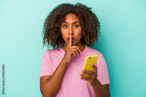 Young african american woman holding a mobile phone isolated on blue background keeping a secret or asking for silence.