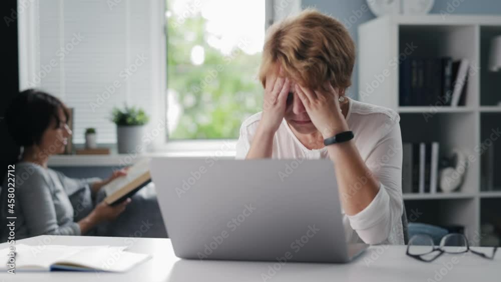 Tired senior woman sitting at table and rubbing eyes after long work on modern laptop. Blur background of her mature daughter relaxing on sofa with book in hands. Tired senior woman sitting at table and rubbing eyes after long work on modern laptop. Blur background of her mature daughter relaxing on sofa with book in hands.