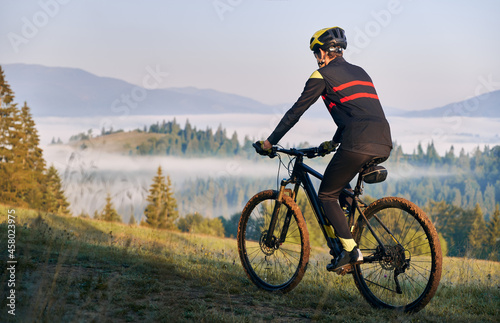 Fototapeta Naklejka Na Ścianę i Meble -  Back view of male cyclist in cycling suit riding bike on road. Man bicyclist wearing safety helmet while enjoying bicycle ride in mountains in the morning. Concept of sport, biking and active leisure.