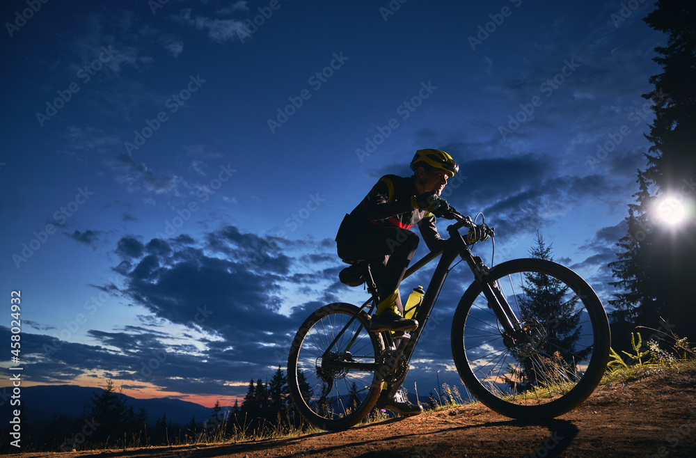 Young man cycling bicycle under beautiful night sky. Male bicyclist in safety helmet riding on hillside road under blue cloudy sky at night. Concept of sport, biking and active leisure.
