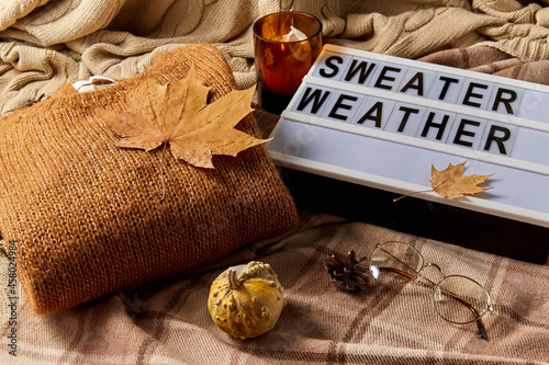 season and objects concept - wool clothes, autumn leaves, glasses and light box with sweater weather letters on warm blankets