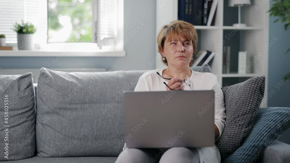 Pensive mature woman in eyeglasses sitting on couch with modern laptop on knees and looking aside. Caucasian lady resting at home alone with her thoughts.