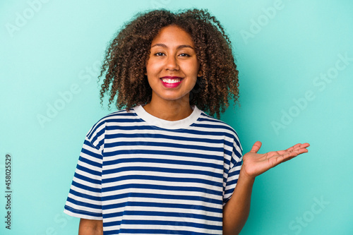 Young african american woman with curly hair isolated on blue background showing a copy space on a palm and holding another hand on waist.