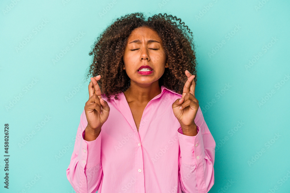 Young african american woman with curly hair isolated on blue background crossing fingers for having luck