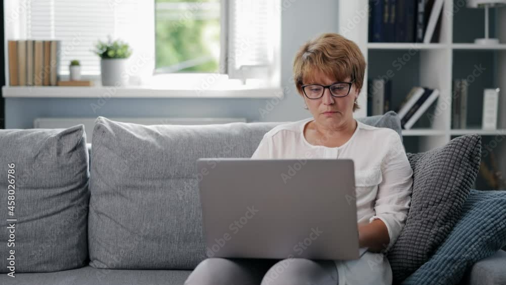 Caucasian female freelancer sitting on couch at living room and typing on modern laptop. Mature woman in eyeglasses working remotely at home. Caucasian female freelancer sitting on couch at living room and typing on modern laptop. Mature woman in eyeglasses working remotely at home.