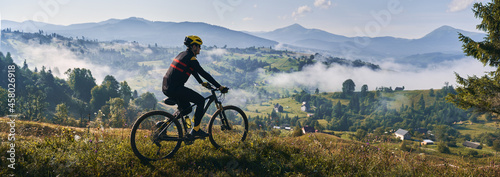 Man riding bicycle on grassy hill and looking at beautiful misty mountains. Male bicyclist enjoying panoramic view of majestic mountains during bicycle ride. Concept of sport, bicycling and nature.