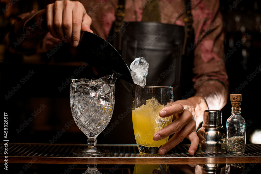 Crystal goblet with ice cubes and glass with drink on bar counter. Hand of bartender pours ice into glass using scoop.