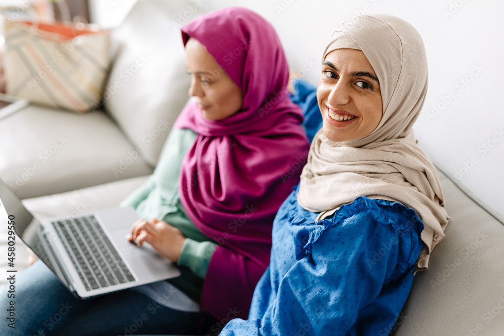Two women in headscarf using laptop while sitting on sofa at home